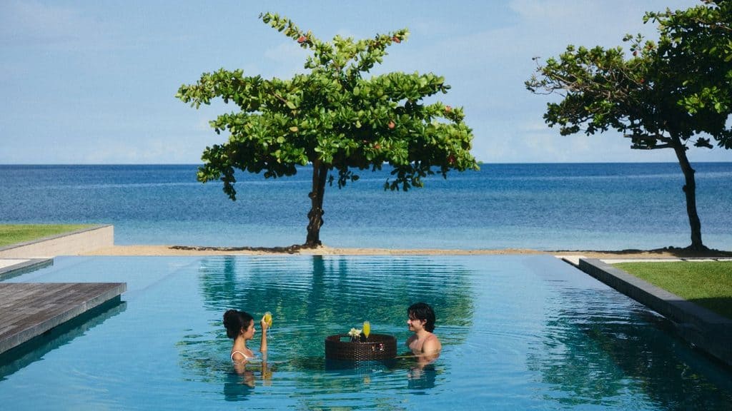 Two people stand in a pool with a floating tray of drinks and food, facing each other, with trees, sandy beach, and ocean in the background.