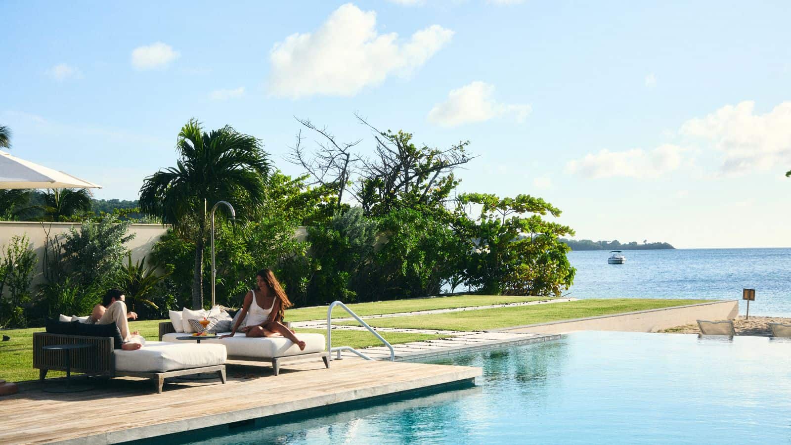 Two people relax on poolside loungers by an infinity pool, with trees, lawn, and a calm ocean in the background under a clear sky.