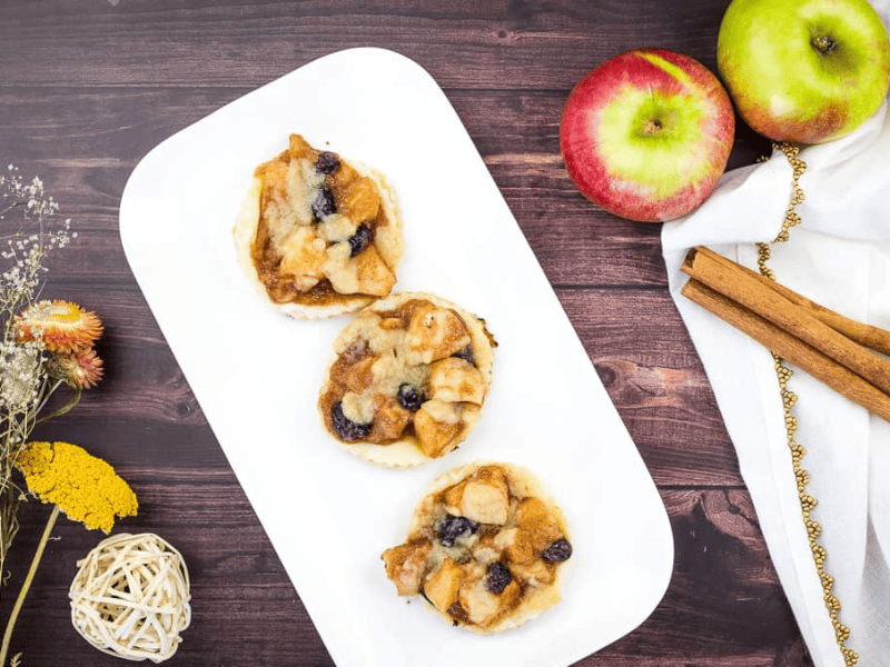 Three small apple and raisin tarts are arranged on a white rectangular plate, next to fresh apples, cinnamon sticks, dried flowers, and a decorative ball on a dark wood surface.