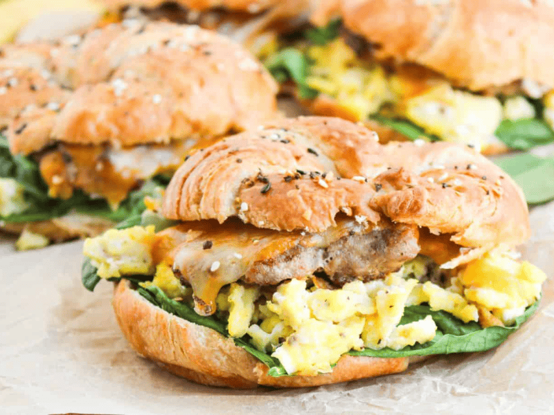 A croissant sandwich filled with scrambled eggs, sausage, cheddar cheese, and spinach on parchment paper.