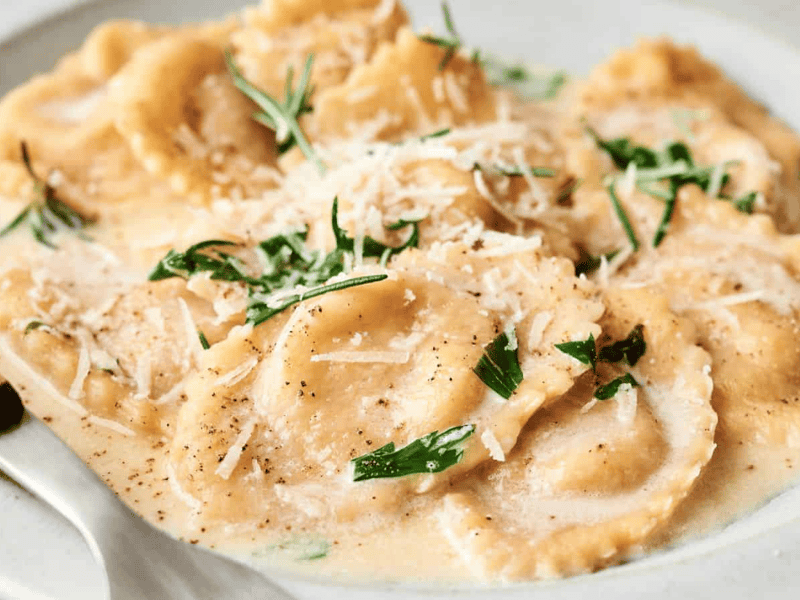 A plate of ravioli topped with creamy sauce, grated cheese, and fresh herbs, served with a fork.