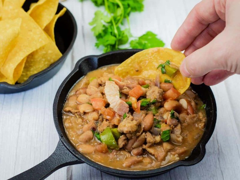 A hand dips a tortilla chip into a small cast-iron skillet filled with Charro Beans With Chorizo, more chips and cilantro are in the background.