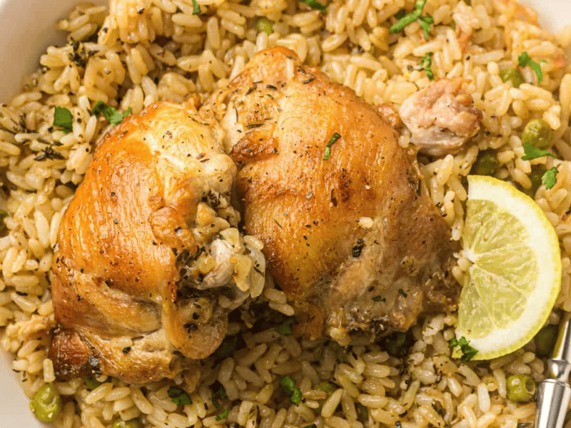 A plate of seasoned baked chicken served on a bed of rice with peas, garnished with chopped herbs and a slice of lemon.