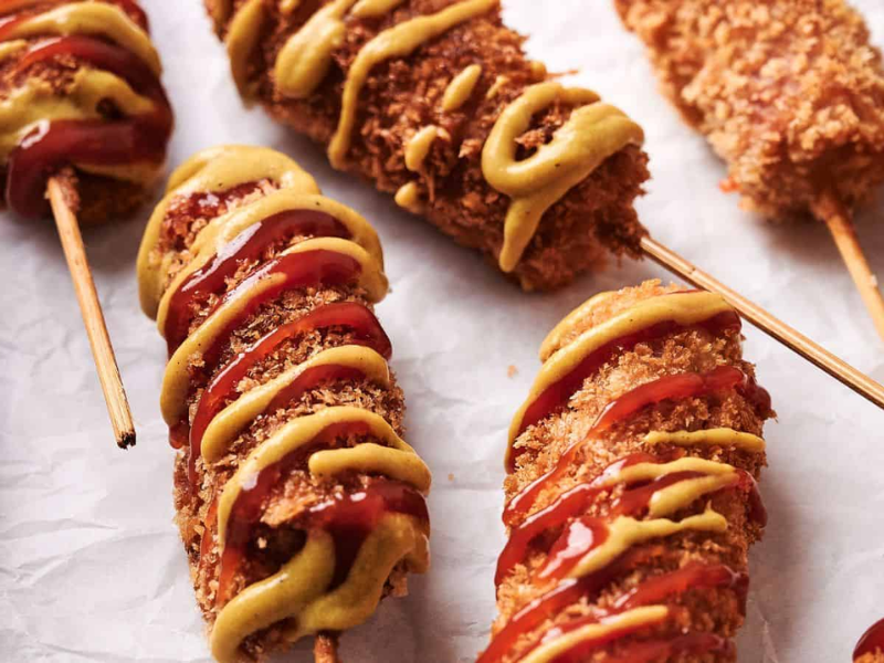 Several breaded and fried corn dogs on sticks, drizzled with mustard and ketchup, placed on white crumpled paper.