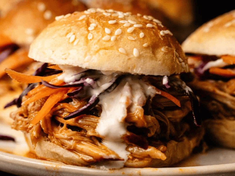 A close-up of a pulled chicken sandwich with sesame seed bun, shredded carrots, cabbage, and creamy sauce.