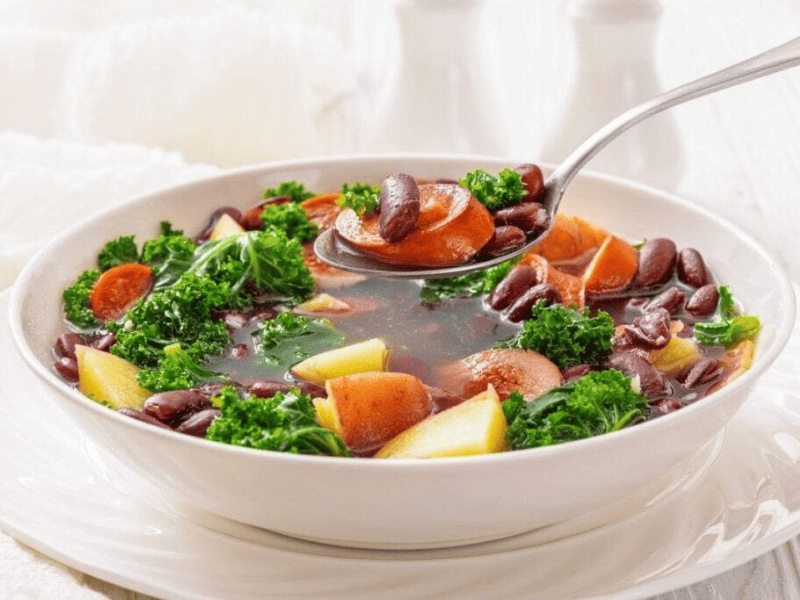 A bowl of soup with red kidney beans, kale, potato chunks, and carrot slices, with a spoon lifting a portion.