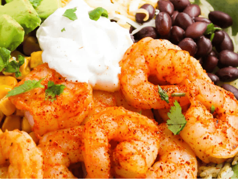 A close-up of a bowl with seasoned shrimp, black beans, avocado, corn, shredded cheese, and a dollop of sour cream, garnished with cilantro.