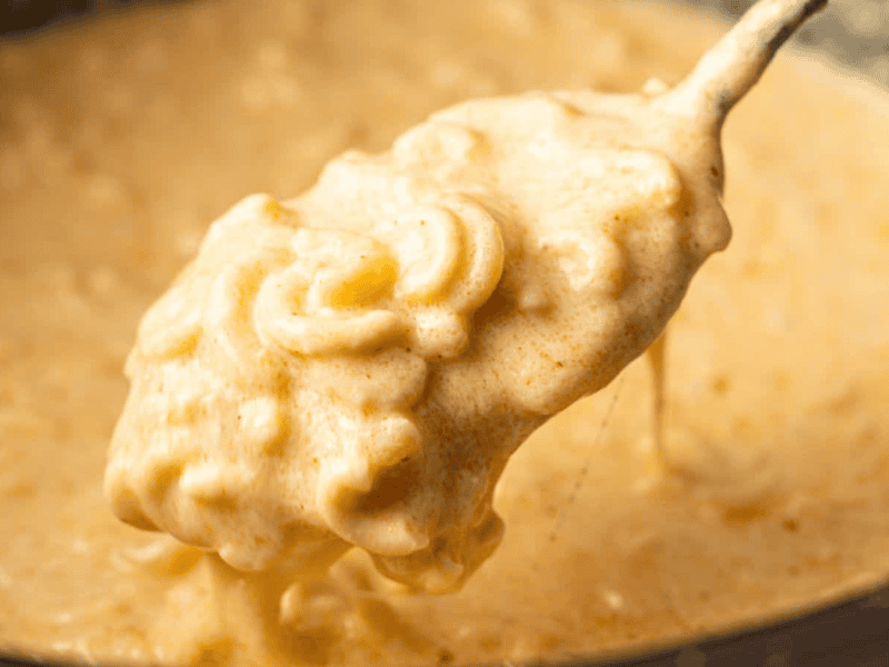 A close-up of a spoon lifting creamy, melted cheese sauce with macaroni from a pot.