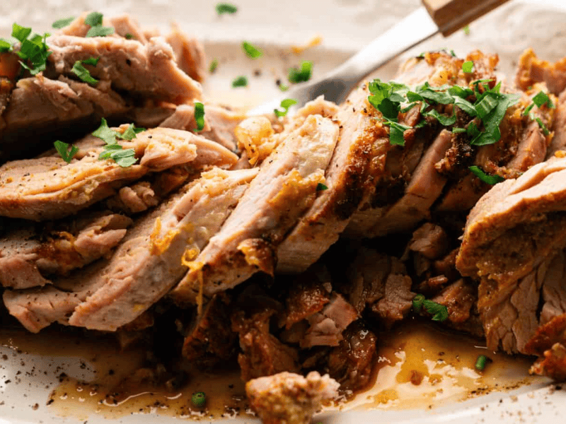 Sliced roasted pork garnished with chopped herbs is arranged on a plate, with visible juices and a serving fork nearby.