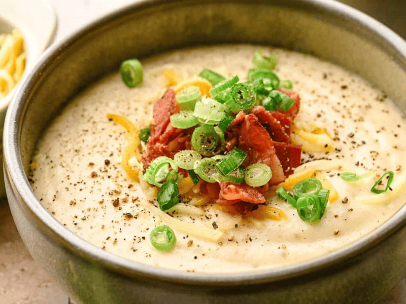 A bowl of creamy soup topped with chopped green onions, grated cheese, crispy bacon pieces, and ground black pepper.