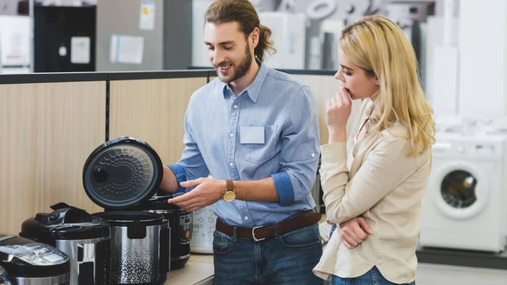 A man shows a multicooker to a woman in an appliance store, with other kitchen appliances and washing machines visible in the background, as slow cookers make a comeback among modern kitchen essentials.