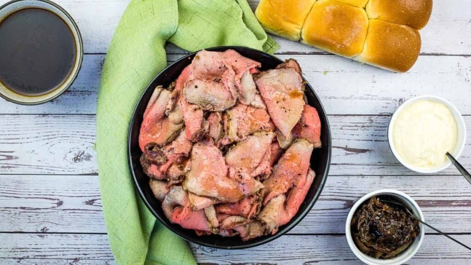 A bowl of sliced roast beef served with dinner rolls, au jus, creamy horseradish sauce, and caramelized onions.