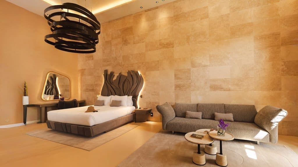 Modern hotel room with a large bed, contemporary headboard, sofa, coffee tables, desk, and a mirror against a tan stone accent wall. Warm lighting enhances the minimalist decor.