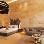 Modern hotel room with a large bed, contemporary headboard, sofa, coffee tables, desk, and a mirror against a tan stone accent wall. Warm lighting enhances the minimalist decor.