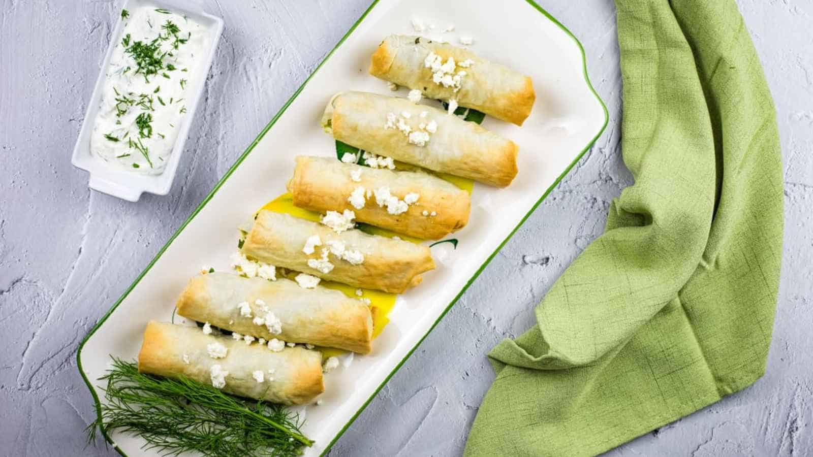 A rectangular platter of cheese-filled phyllo rolls topped with crumbled feta, served with a side of herb-topped yogurt dip.
