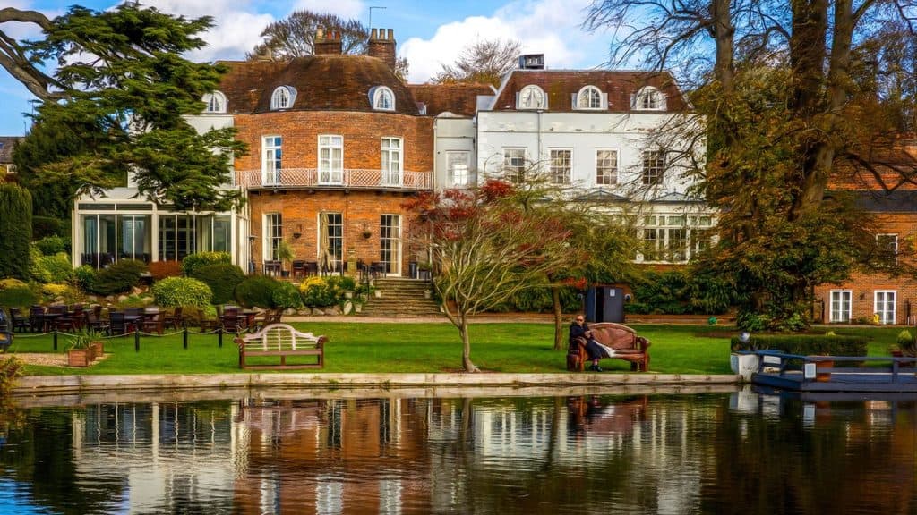 Large brick and white house with multiple windows, a balcony, outdoor seating, well-kept lawn, trees, and a river in the foreground reflecting the building.