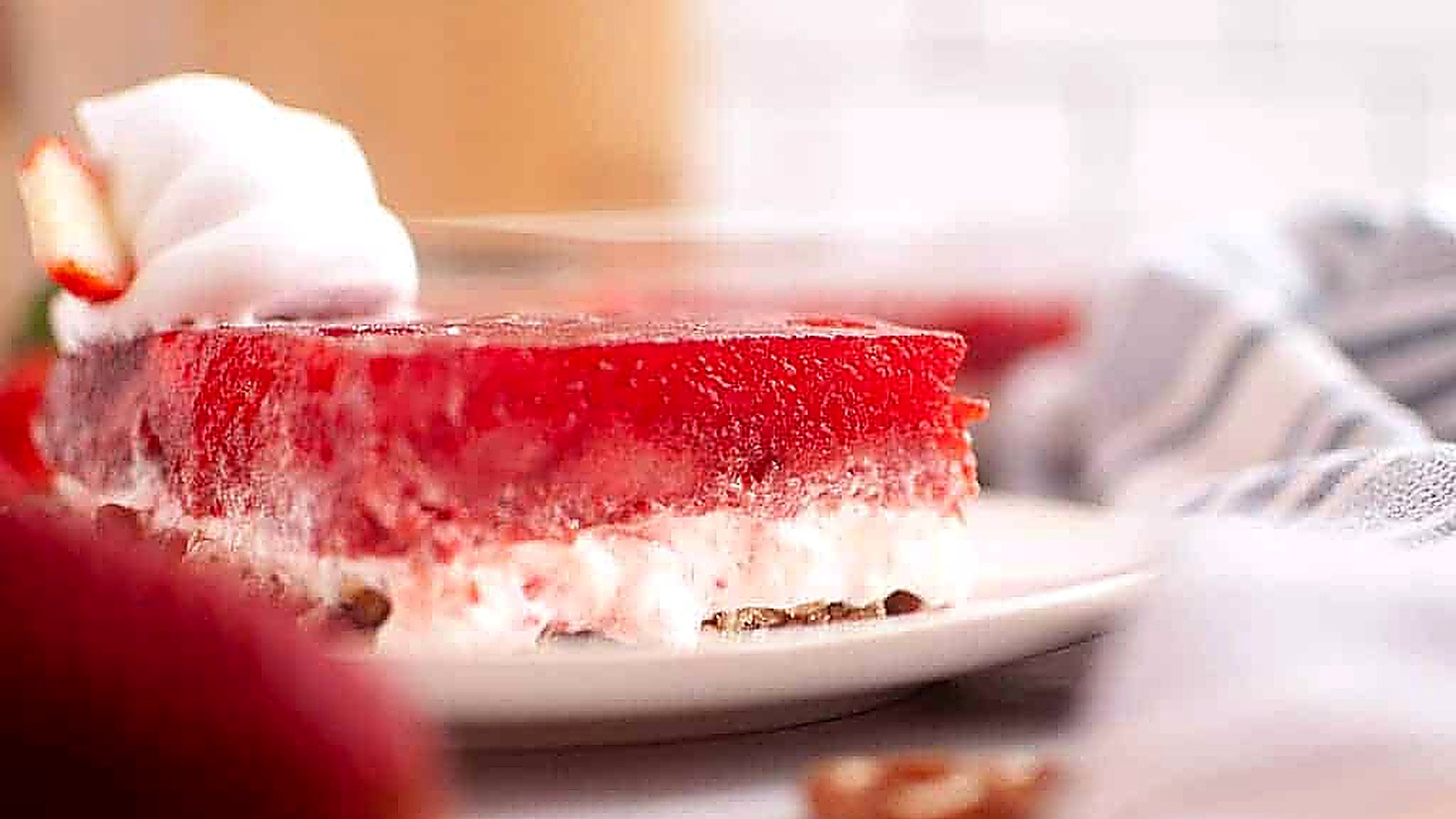 A slice of strawberry pretzel salad with whipped cream on a white plate, showing layers of red gelatin, creamy filling, and pretzel crust.