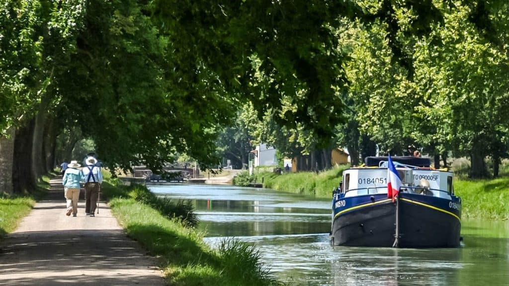 A boat with a French flag traveling along a canal during a Gascony barge cruise, with two people walking along the tree-lined towpath.