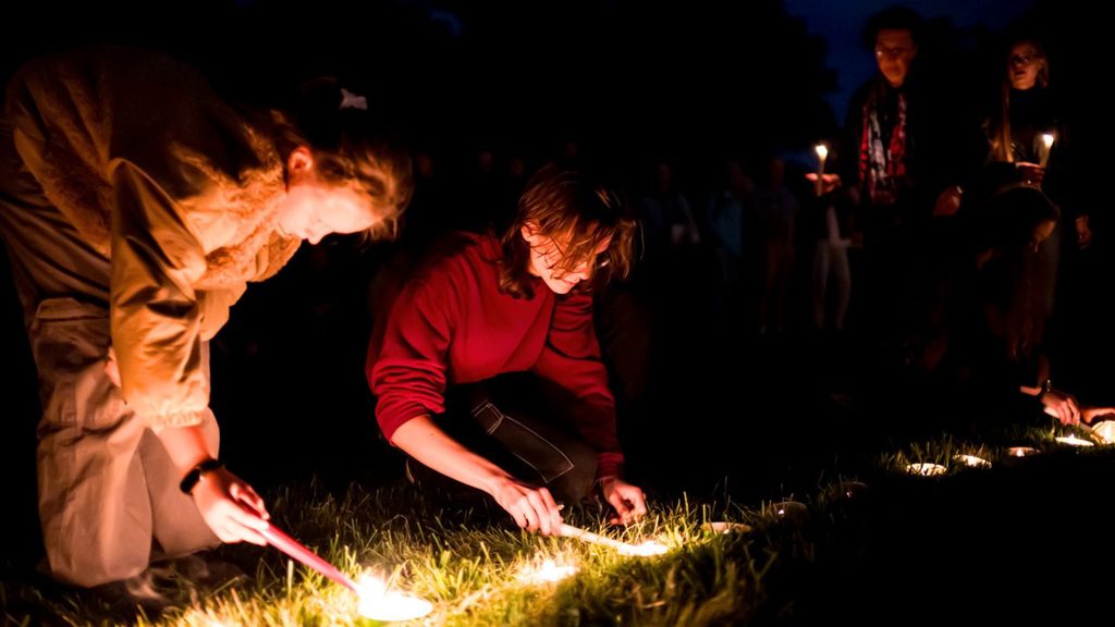 People kneeling on grass light candles at night during a vigil, with others standing in the background holding candles.