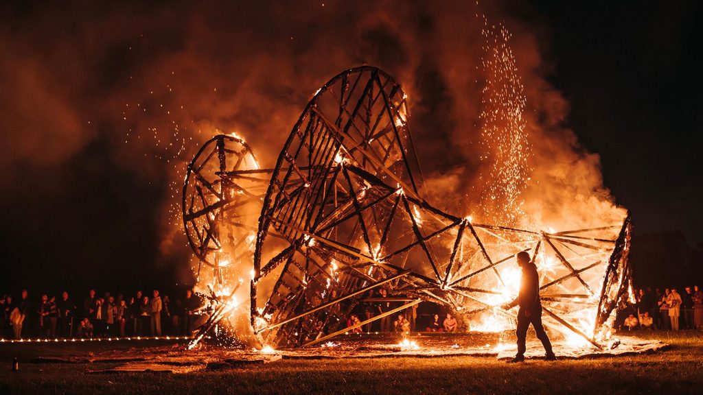 A large wooden sculpture burns at night as a crowd watches from a distance; a person stands nearby, silhouetted by the flames and sparks rise into the dark sky.