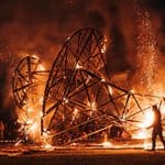A large wooden sculpture burns at night as a crowd watches from a distance; a person stands nearby, silhouetted by the flames and sparks rise into the dark sky.