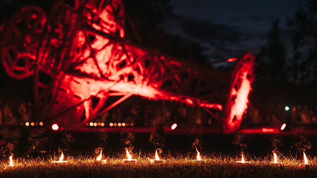 A row of small fires burns on grass at night, with a large red-lit metal structure and dark trees visible in the background.