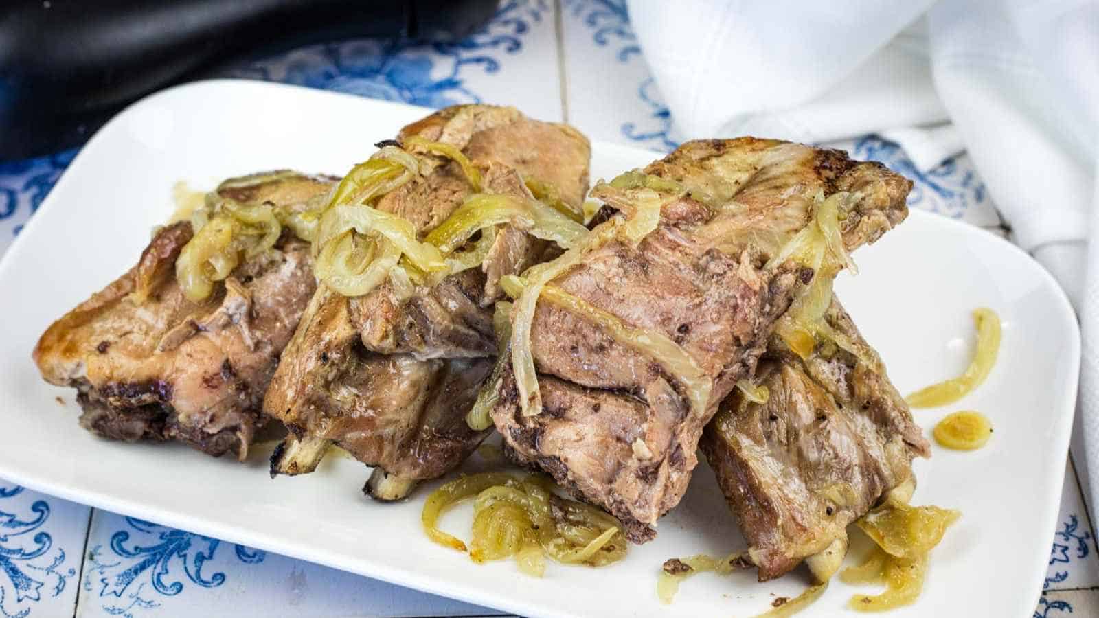 Sunday pork ribs topped with sautéed onions are arranged on a white rectangular plate.