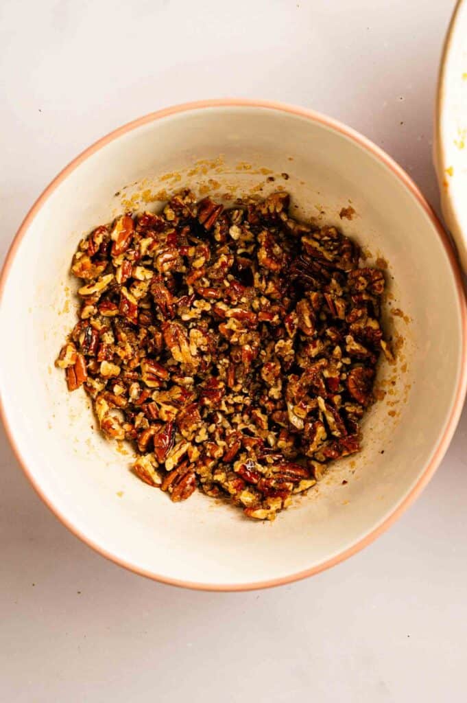 A mixing bowl filled with chopped pecans coated in a sticky brown mixture, placed on a light surface.