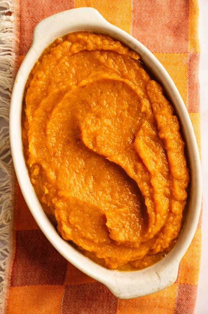 Oval white dish filled with smooth, orange mashed sweet potatoes, placed on an orange and yellow checkered cloth with fringe on the side.