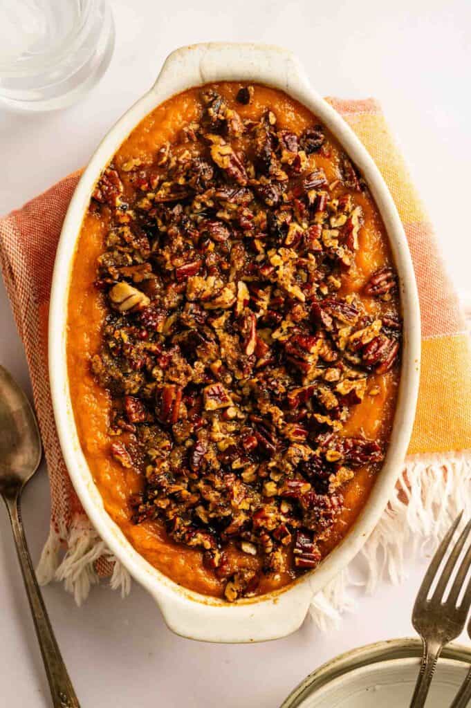 Oval dish of sweet potato casserole topped with a brown pecan crumble, set on an orange napkin with spoons and a glass of water nearby.