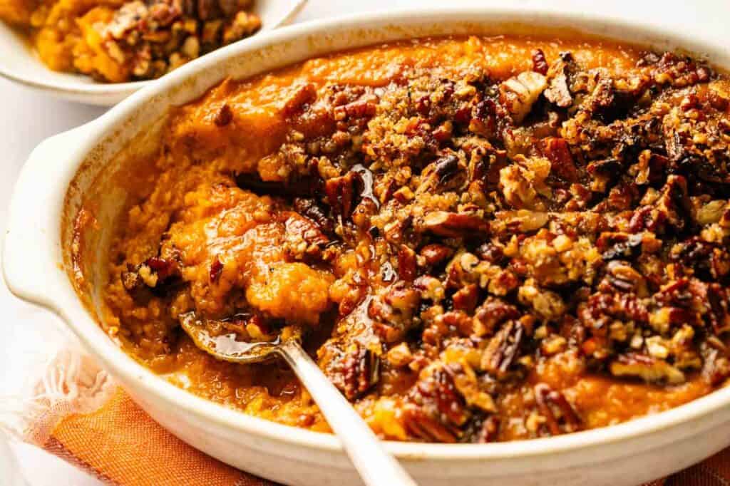 Baked sweet potato casserole topped with chopped pecans in a white dish, with a serving spoon and a small portion scooped out.