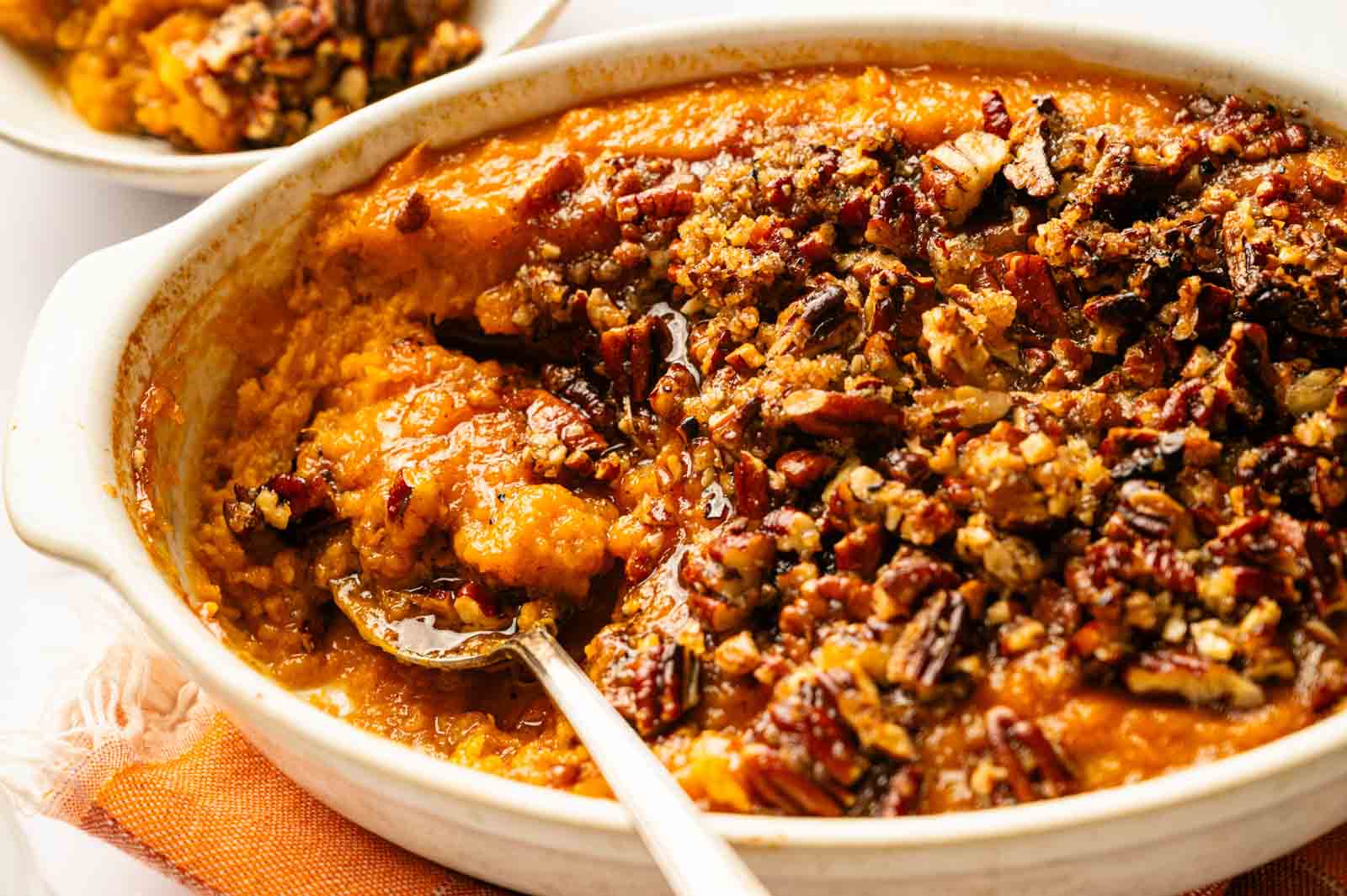 Baked sweet potato casserole topped with chopped pecans in a white dish, with a serving spoon and a small portion scooped out.