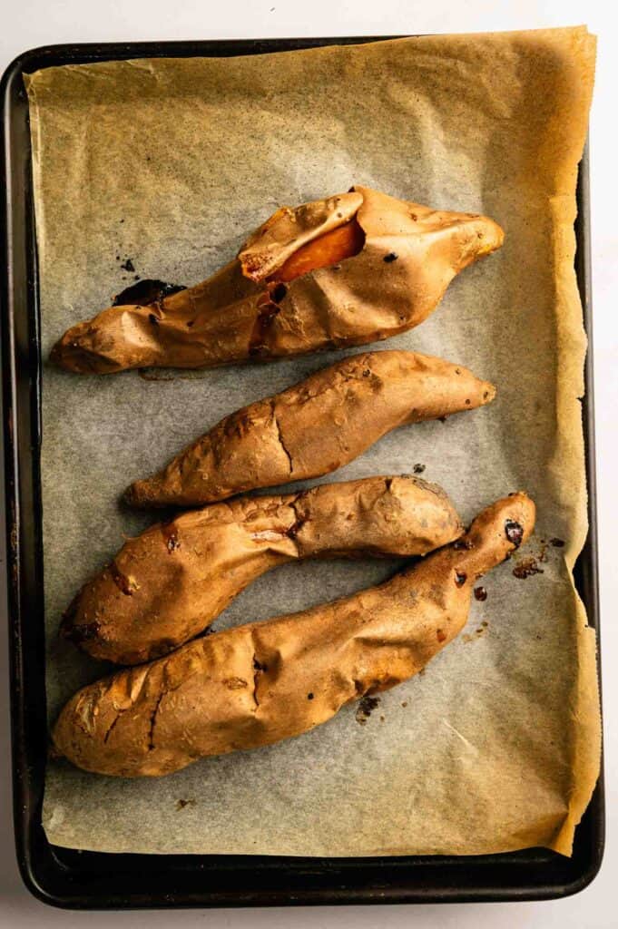 Four baked sweet potatoes with cracked skins on a parchment-lined baking tray, viewed from above.