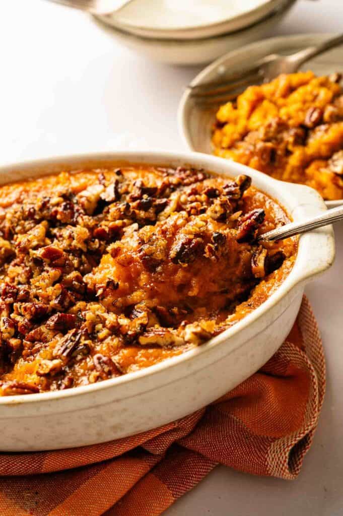 A casserole dish filled with sweet potato casserole topped with chopped pecans, with a serving spoon lifting out a portion.