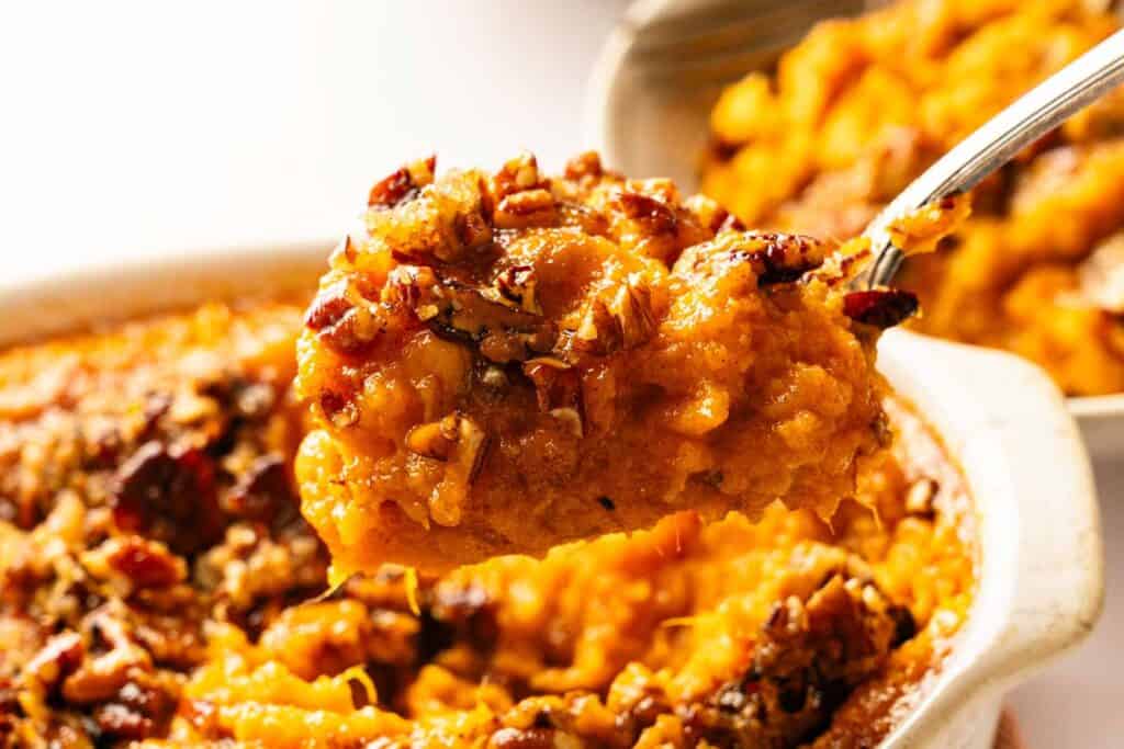 A close-up of a spoonful of sweet potato casserole topped with chopped pecans, held above a baking dish containing more of the casserole.