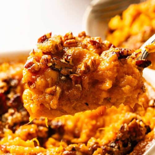 A close-up of a spoonful of sweet potato casserole topped with chopped pecans, held above a baking dish containing more of the casserole.