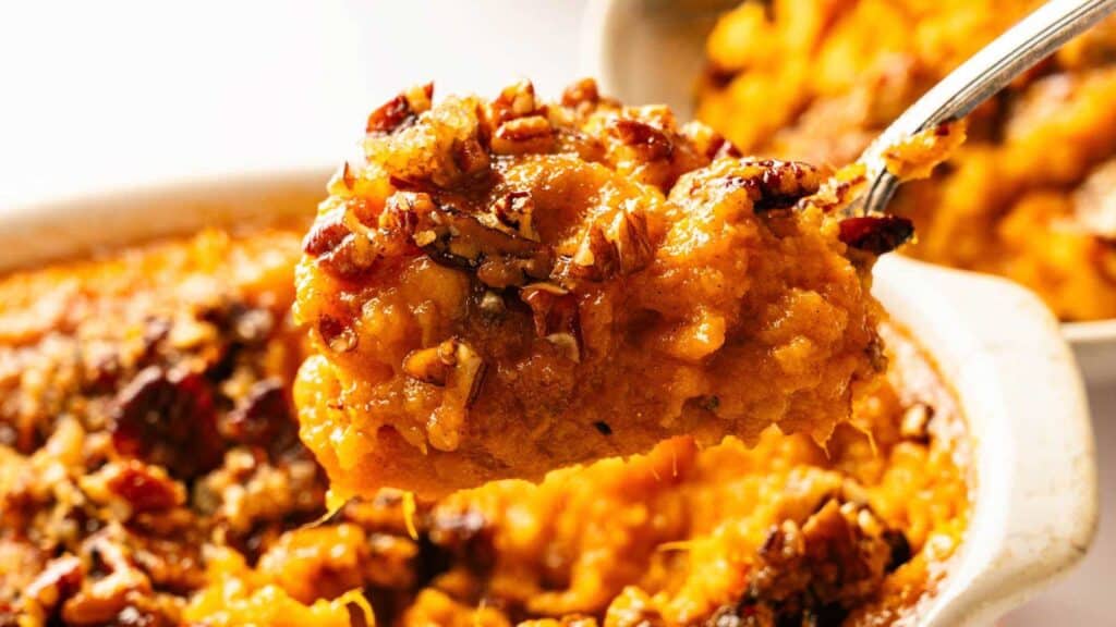 A close-up of a spoonful of sweet potato casserole topped with chopped pecans, lifted from a baking dish.