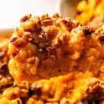 A close-up of a spoonful of sweet potato casserole topped with chopped pecans, lifted from a baking dish.