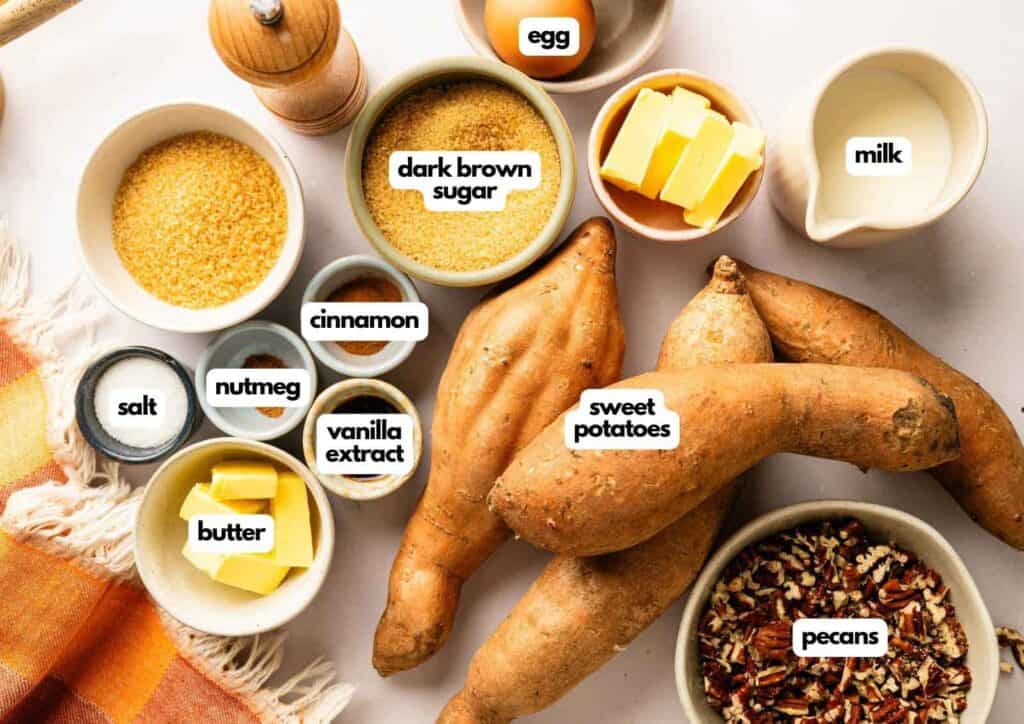 Whole sweet potatoes, bowls of pecans, dark brown sugar, butter, milk, egg, nutmeg, cinnamon, vanilla extract, and salt are arranged on a white surface.
