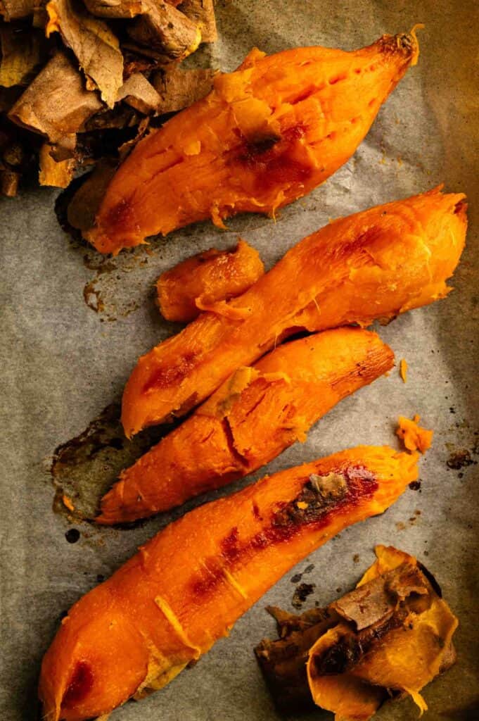 Roasted sweet potatoes with split skins, arranged on parchment paper, with some pieces of peel off to the side.