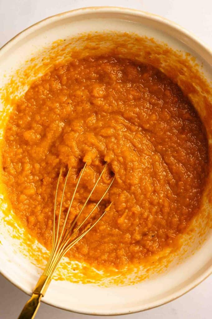 A whisk rests in a bowl filled with a thick, orange mixture, likely pumpkin puree or batter, seen from above.