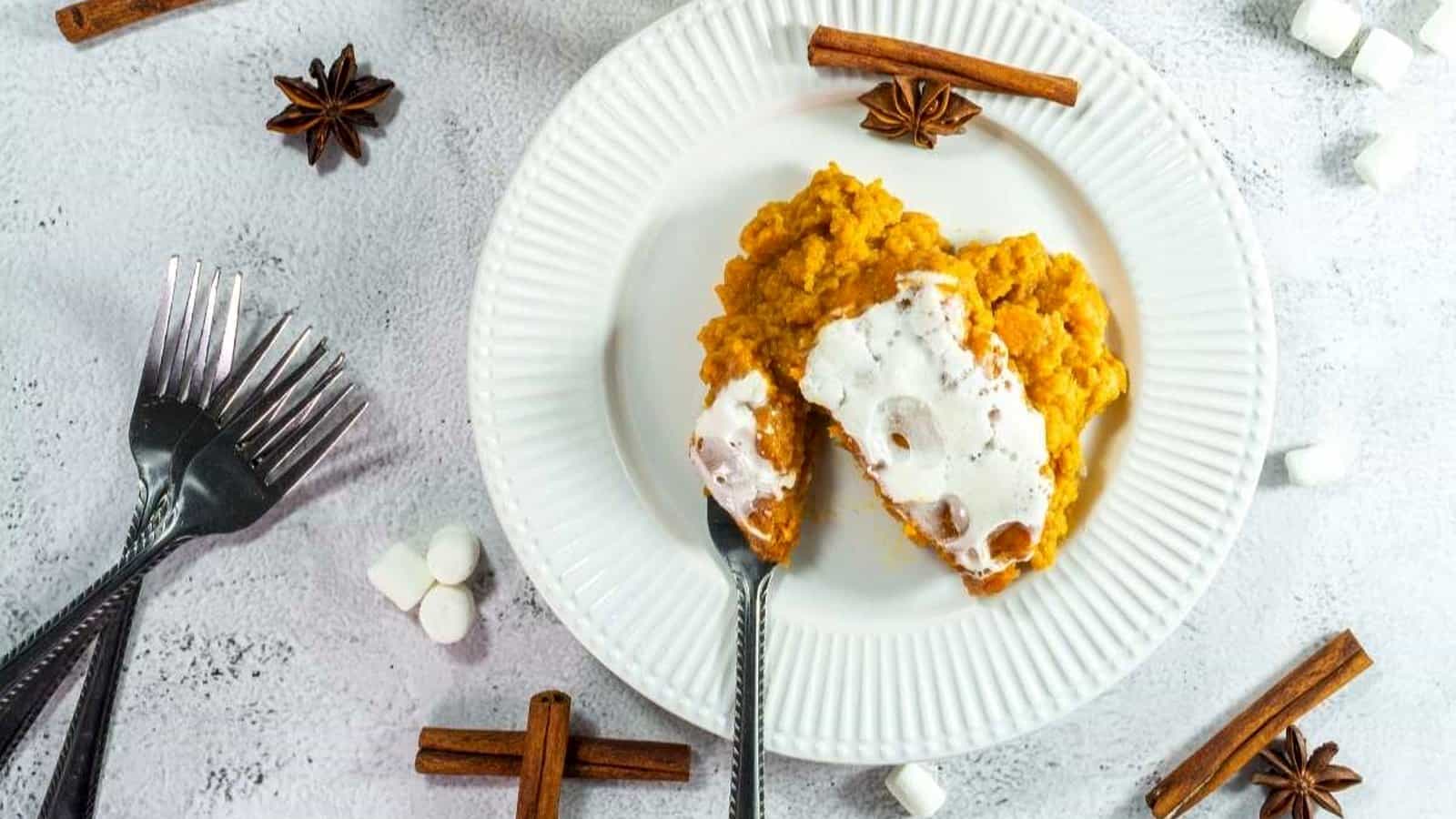 A plate with a serving of sweet potato casserole topped with marshmallows, surrounded by forks, marshmallows, cinnamon sticks, and star anise on a white surface.