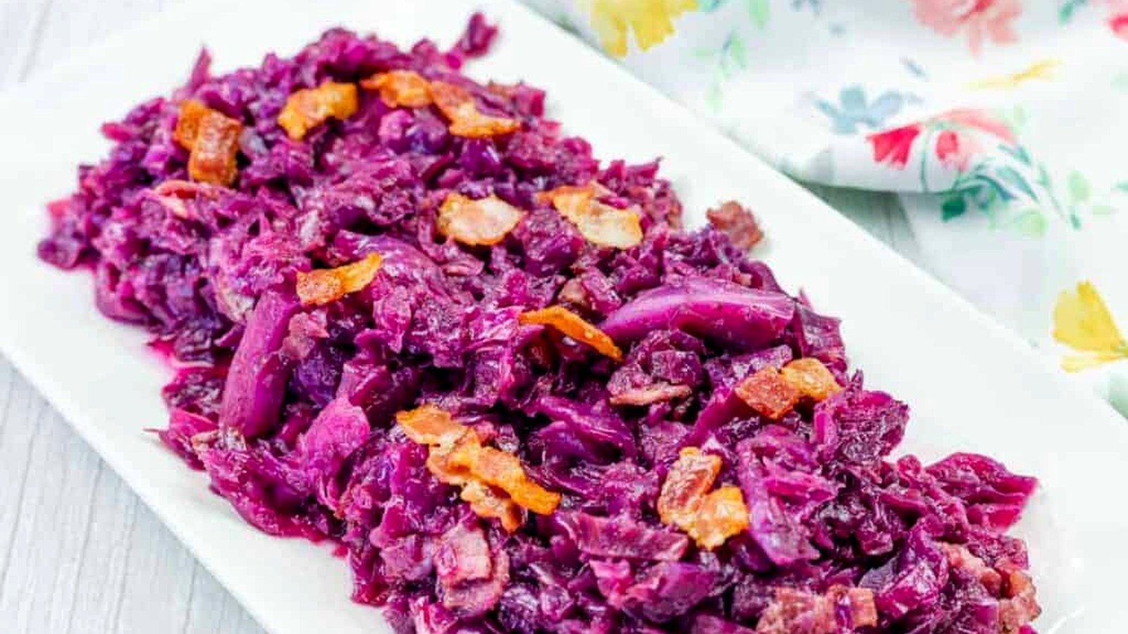 A rectangular white plate with cooked red cabbage and bacon pieces, placed on a light surface next to a floral-patterned cloth.