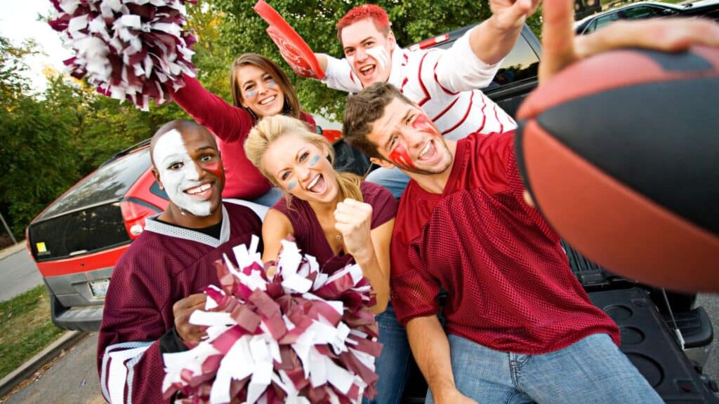 Five people with face paint and sports attire pose enthusiastically with pom-poms and a football near parked cars, likely at a tailgate event.