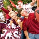 Five people with face paint and sports attire pose enthusiastically with pom-poms and a football near parked cars, likely at a tailgate event.