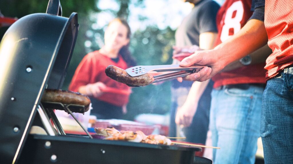 Person grilling sausages and skewers on a barbecue while others stand nearby outdoors, with one person wearing a red sports jersey.
