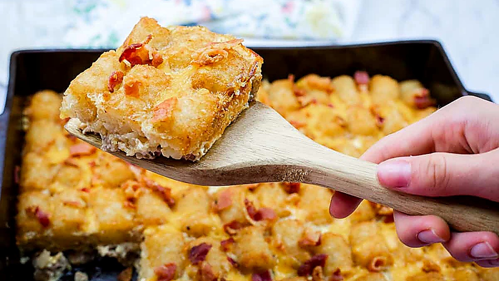 A wooden spatula holds a square piece of Crispy Tater Tot Breakfast Bake with bits of bacon, with the rest of the casserole visible in the background.