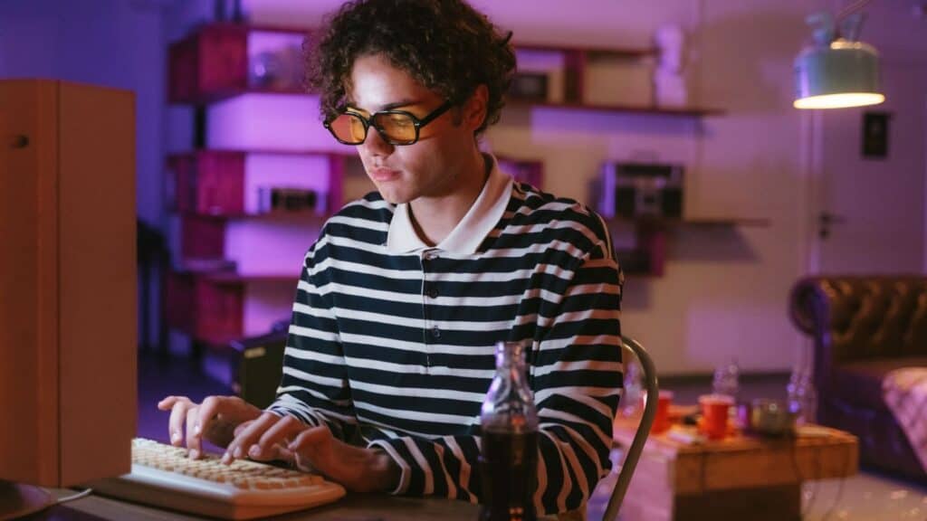 Person wearing a striped shirt and yellow glasses types on a computer keyboard at a desk with a drink, in a dimly lit room with shelves and a couch in the background&mdash;a nostalgic scene reminiscent of 90s kids logging onto dial up internet.