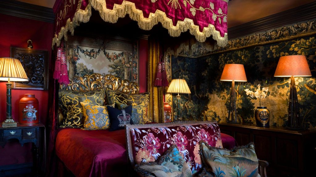 Opulent bedroom with a red canopy bed, ornate pillows, dark wood furniture, patterned wallpaper, and warm lamp lighting.