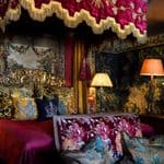 Opulent bedroom with a red canopy bed, ornate pillows, dark wood furniture, patterned wallpaper, and warm lamp lighting.